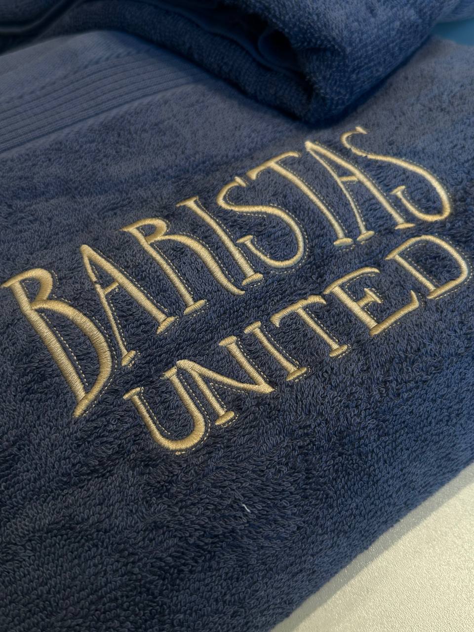 Firmenkleidung besticken Wien: dunkelblaues Frottee Handtuch mit goldener Baristas United Logostickerei